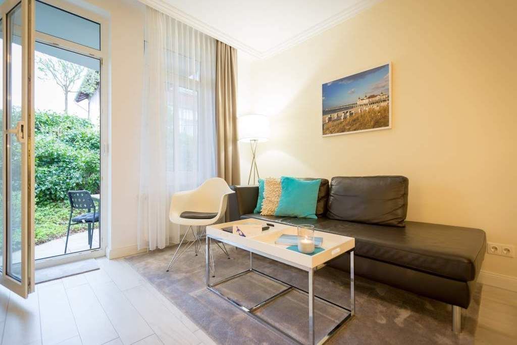 Una sala de estar moderna con un sofá, una mesa de centro y una silla. Hay un ventanal con cortinas, un cuadro en la pared y vistas a zonas verdes.