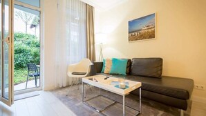Een moderne woonkamer met een bank, een salontafel en een stoel. Er is een groot raam met gordijnen, een schilderij aan de muur en uitzicht op groen buiten.