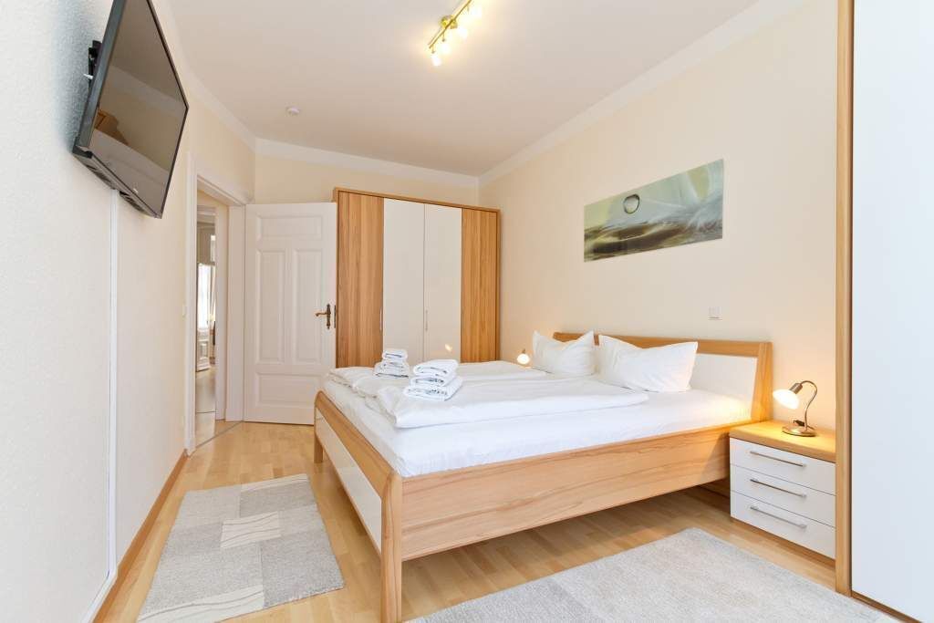 A bedroom with a wooden bed, white bedding, a bedside table with a lamp, and a TV mounted on the wall.