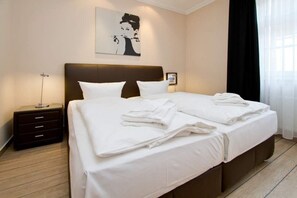 Una cama bien hecha con sábanas y toallas blancas, un cabecero oscuro, una mesita de noche con una lámpara y un cuadro en la pared.