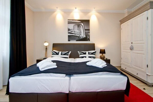 Una cama doble con una colcha blanco y negro, dos almohadas con diseños de la bandera Union Jack, una manta roja y negra y una toalla blanca doblada sobre la cama.