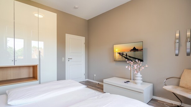 Een slaapkamer met een bed, nachtkastje, stoel, kledingkast en een aan de muur bevestigde televisie waarop een zonsondergang te zien is.
