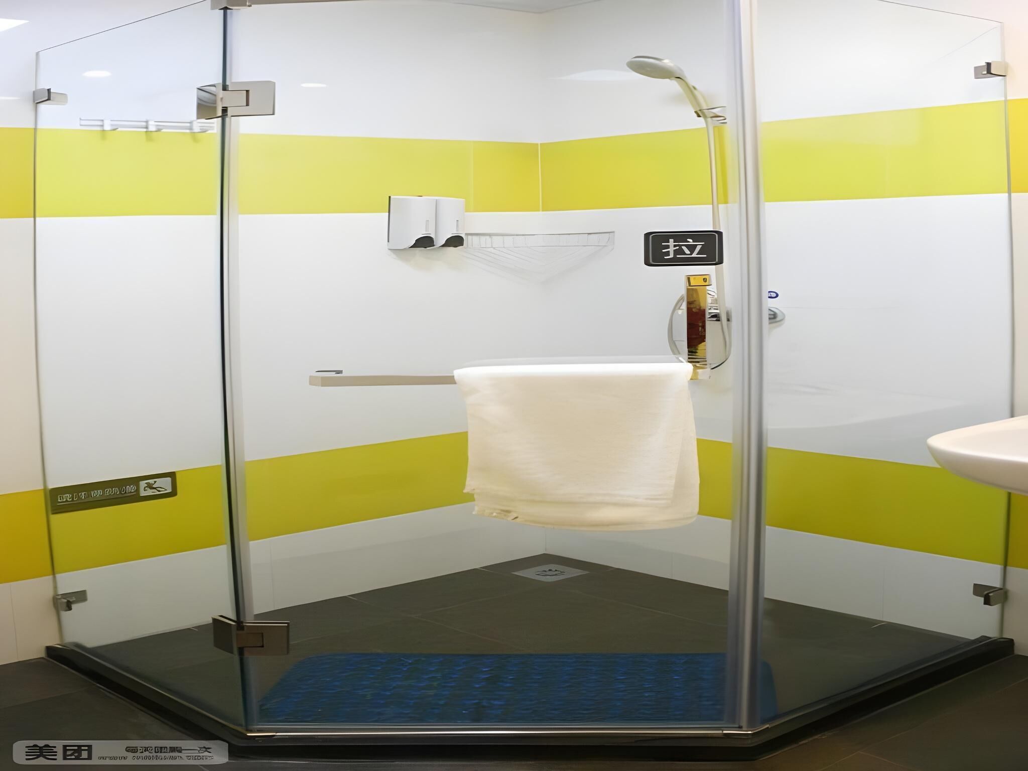 A glass-enclosed shower stall with a white towel hanging on a rack, a handheld showerhead, and a soap dispenser on the wall.
