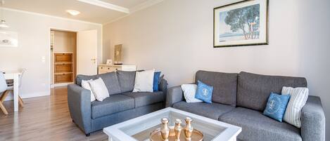 Ein modernes Wohnzimmer mit einem grauen Ecksofa, einem Glastisch und einem Essbereich mit weißem Tisch und Stühlen.