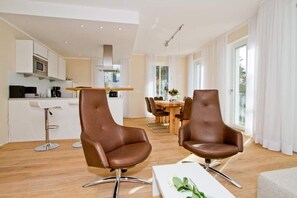 Una cocina moderna con armarios blancos y electrodomésticos de acero inoxidable, un comedor con mesa de madera y sillas de cuero marrón, y ventanas amplias con cortinas blancas.