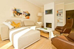Un salon moderne avec un canapé, un pouf, une table basse et un fauteuil en cuir marron. Les murs sont ornés de tableaux abstraits et une bibliothèque remplie de livres se trouve dans la pièce.