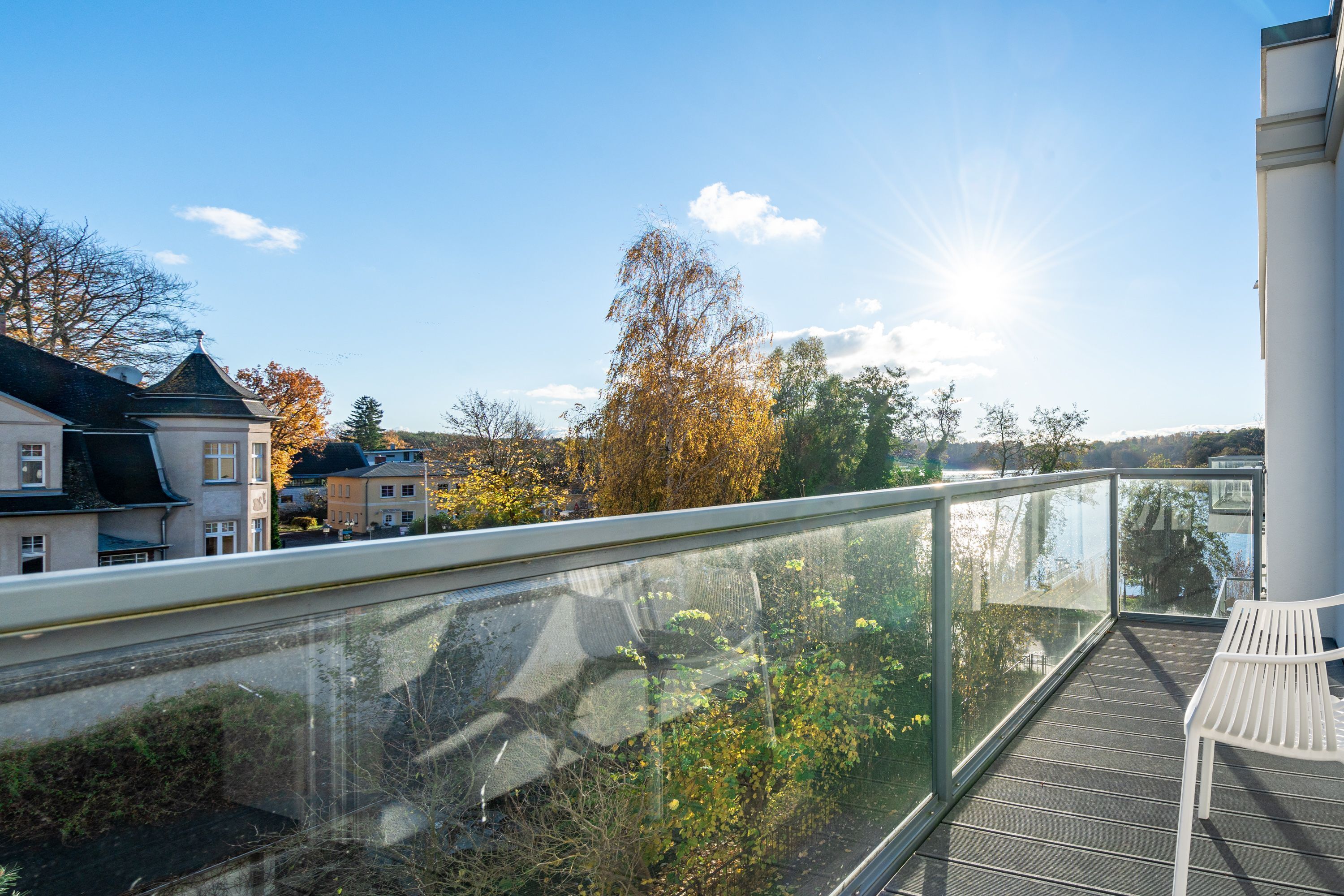 A balcony with a glass railing overlooking a residential area, a white chair, and a clear sky.