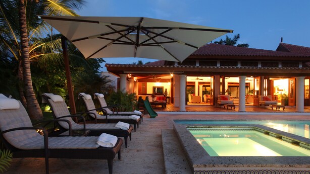 Zona junto a la piscina con sillones de descanso y sombrillas, piscina y un edificio con terraza abierta.