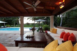 Ein Außenbereich mit Terrasse, Pool, Holzboden, einem weißen Sofa mit roten und gelben Kissen sowie einem Tisch mit einer Vase und Kerzen.