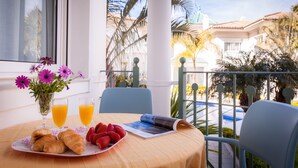 A table with croissants, strawberries, and juice glasses, a vase with flowers, and a magazine on a balcony overlooking palm trees and a pool. - Marina Turquesa - Apartment 2 bedrooms 2 bathrooms and terrace garden views (Nerja)
