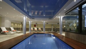 An indoor swimming pool with clear blue water, surrounded by white lounge chairs with orange cushions, and a modern architectural design with glass walls and a high ceiling. - Marina Turquesa - Apartment 2 bedrooms 2 bathrooms and terrace garden views (Nerja)