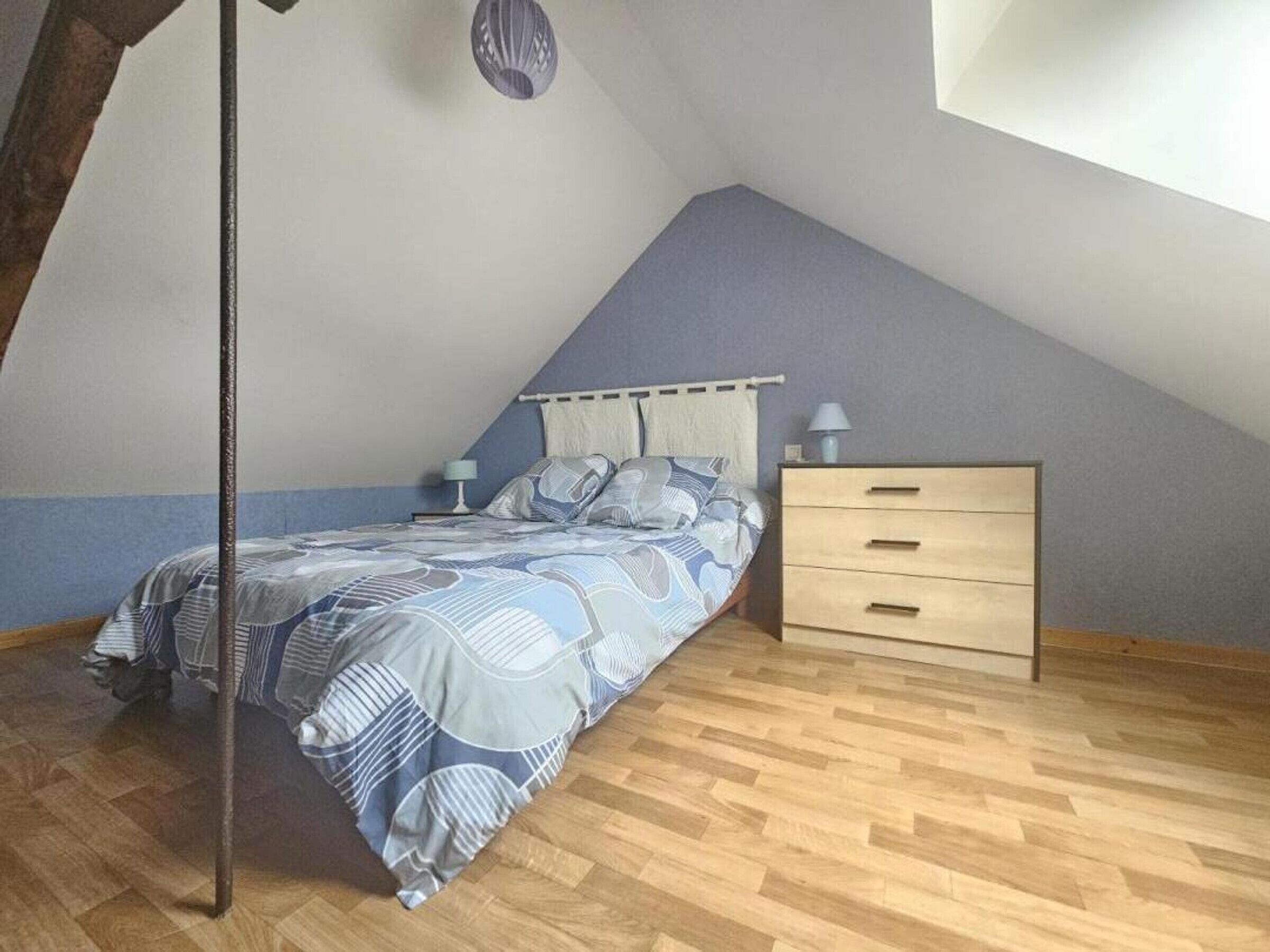 Een slaapkamer met een schuine plafond, een bed met beddengoed met een patroon, een nachtkastje met een lamp, en een houten dressoir.