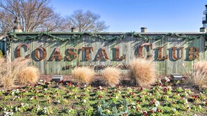 Une enseigne en bois sur laquelle on peut lire en grandes lettres « Coastal Club », entourée de plantes décoratives et se dressant sous un ciel d’un bleu limpide.