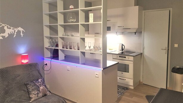A compact living space with a kitchenette, a sofa with a patterned cushion, and a wall-mounted shelf with various glassware. - Cosy 2-Room Apartment with Parking, Private Ski Locker, and Modern Amenities in Bolquère (Bolquère-Superbolquère)
