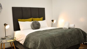 A neatly made bed with a dark headboard, white and mustard pillows, and a grey accent pillow. There are two bedside lamps and a small table with a glass top.