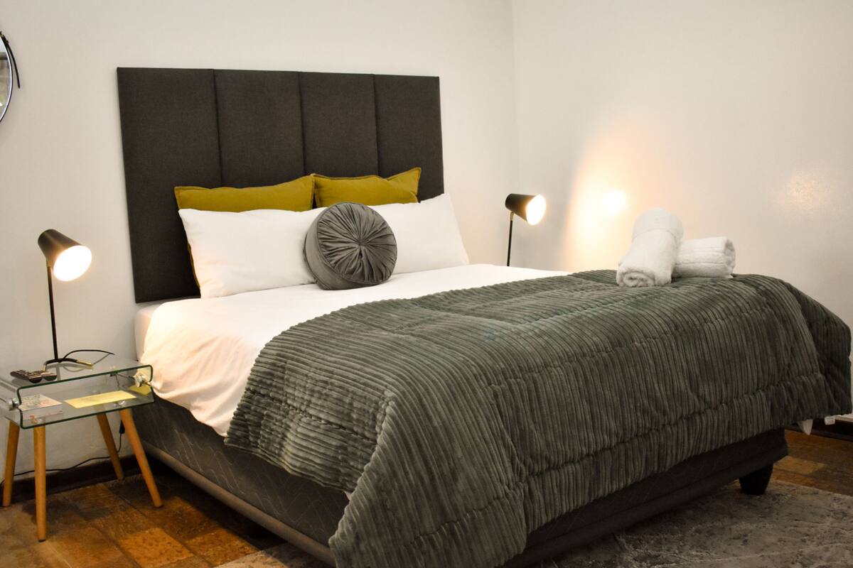 A neatly made bed with a dark headboard, white and mustard pillows, and a grey accent pillow. There are two bedside lamps and a small table with a glass top.