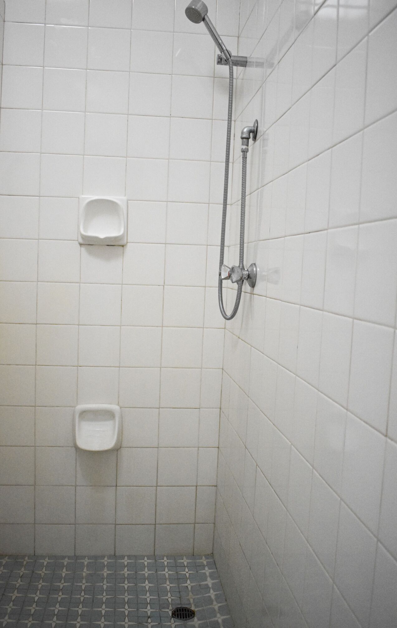 Un petit espace de douche carrelé, équipé d’un pommeau de douche à main et de deux urinoirs fixés au mur.