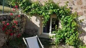A stone building with a small window and a white door, surrounded by greenery and red flowers.