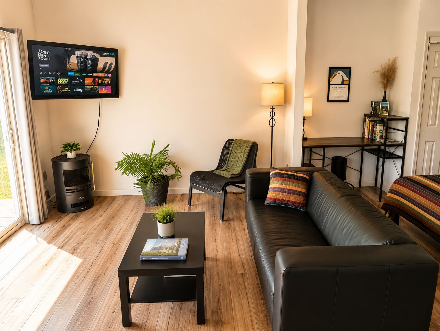 Una sala de estar con un sofá de cuero negro, una mesa de centro negra con un libro, una planta en maceta, un televisor montado en la pared mostrando Dove Men + Care, y un escritorio con una lámpara.