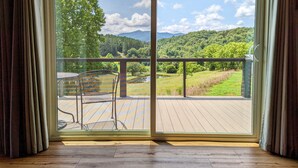 Una habitación con un gran ventanal corredero que da a una terraza exterior con una silla metálica y vistas a un paisaje verde con árboles y colinas.