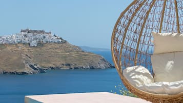Una silla colgante de mimbre con un cojín, un libro, un vaso de zumo de naranja y un sombrero de paja sobre una mesa junto a una piscina con vistas a un pueblo costero y un castillo.