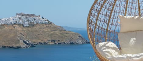 A wicker hanging chair with a cushion and a book, a glass of orange juice, and a straw hat on a table by a pool with a view of a coastal town and a castle.