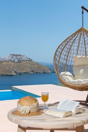 Una silla colgante de mimbre con un cojín, un libro, un vaso de zumo de naranja y un sombrero de paja sobre una mesa junto a una piscina con vistas a un pueblo costero y un castillo.