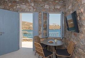 A room with stone walls, two open windows with a view of a coastal landscape, a round table with two chairs, and a flat-screen TV mounted on the wall. - Nisides Suites (Astypalaia)