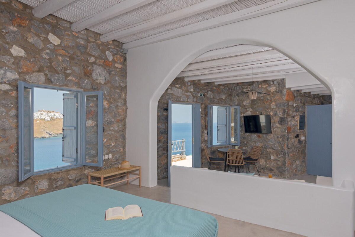 A bedroom with a stone wall, a bed with a blue bedspread, and a view of the sea through open windows.