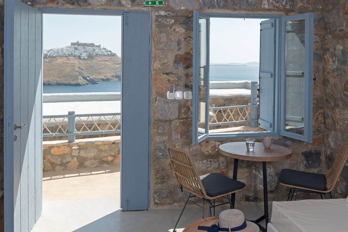A room with stone walls, a view of the sea, a table with chairs, and a hanging blue light fixture.