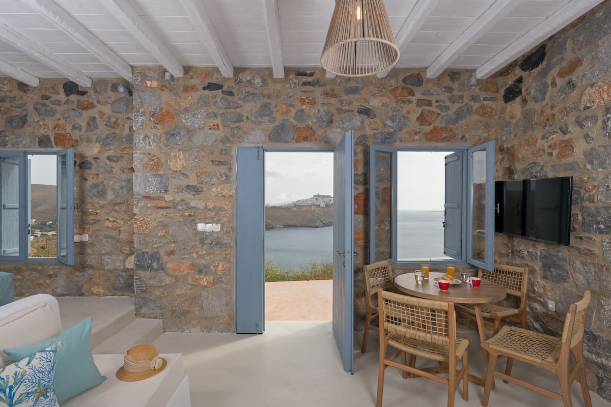 A stone-walled room with a dining area, a television, and a view of the sea.