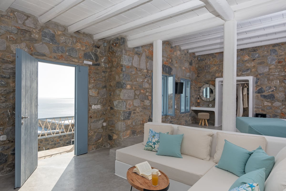 A stone-walled room with a white sofa, a small round table with a book and glass, and a view of the sea through an open door.