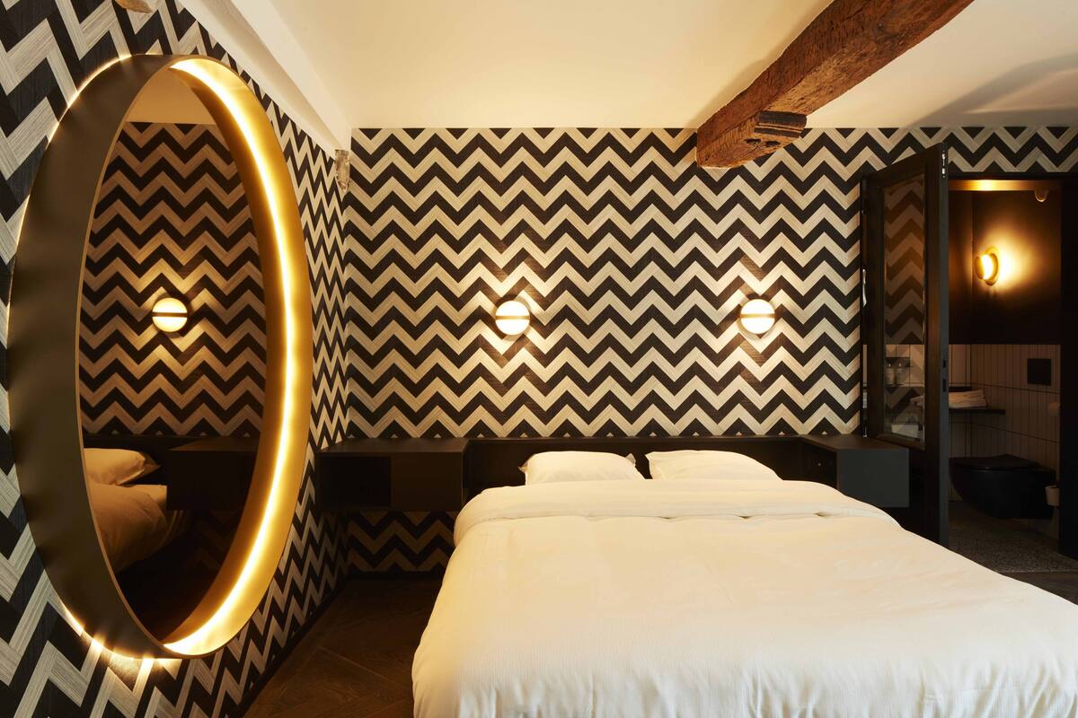 A modern hotel room with a large bed, a circular mirror, and a chevron-patterned wall.
