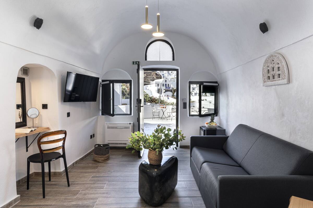 A compact living space with a sofa, a chair, a small table, a TV mounted on the wall, and a view of the outside through a window.