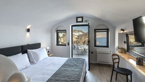 A bedroom with a bed, a desk, a chair, and a TV. There are windows with views of the outside. - Pearl of Caldera Oia Boutique Hotel (Santorini)