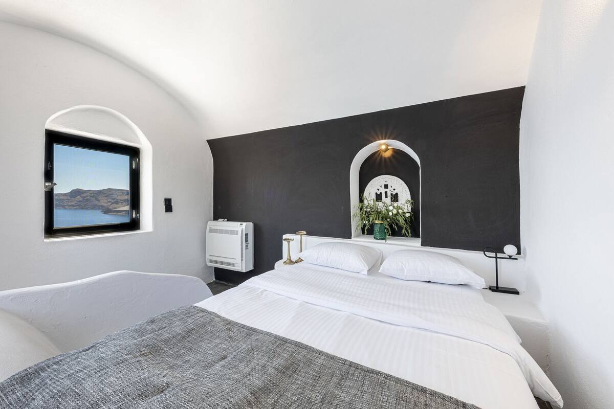 A modern bedroom with a large bed, a window with a view of the sea, a wall-mounted air conditioner, and a decorative mirror.