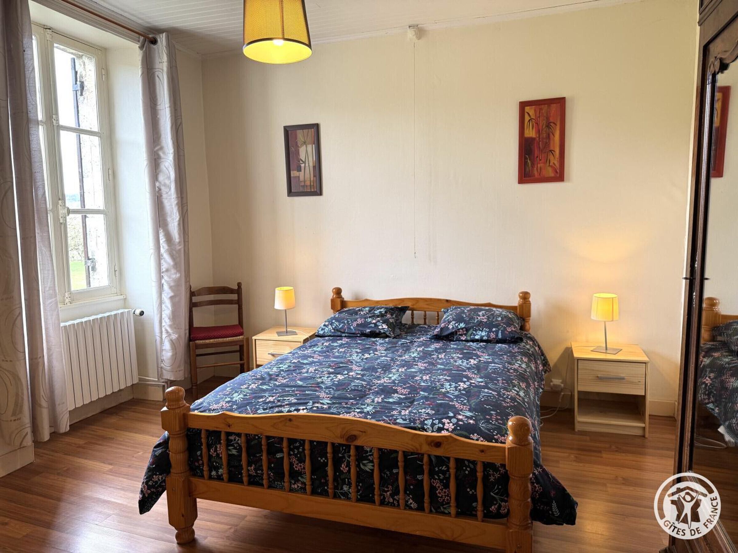 Une chambre à coucher avec un lit en bois, deux tables de chevet avec des lampes, un radiateur, une fenêtre avec des rideaux et deux tableaux accrochés au mur.