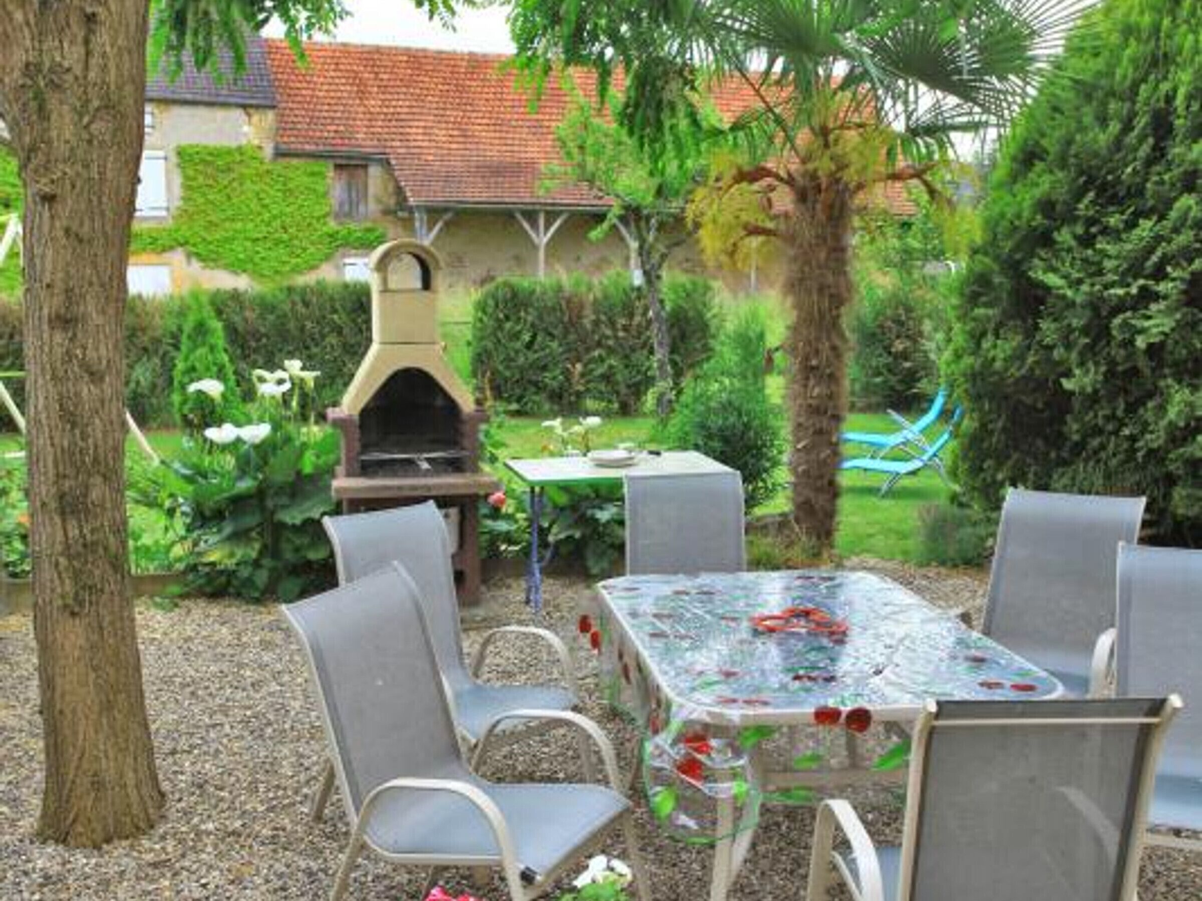 An outdoor patio with a table and chairs, surrounded by greenery and a barbecue grill.