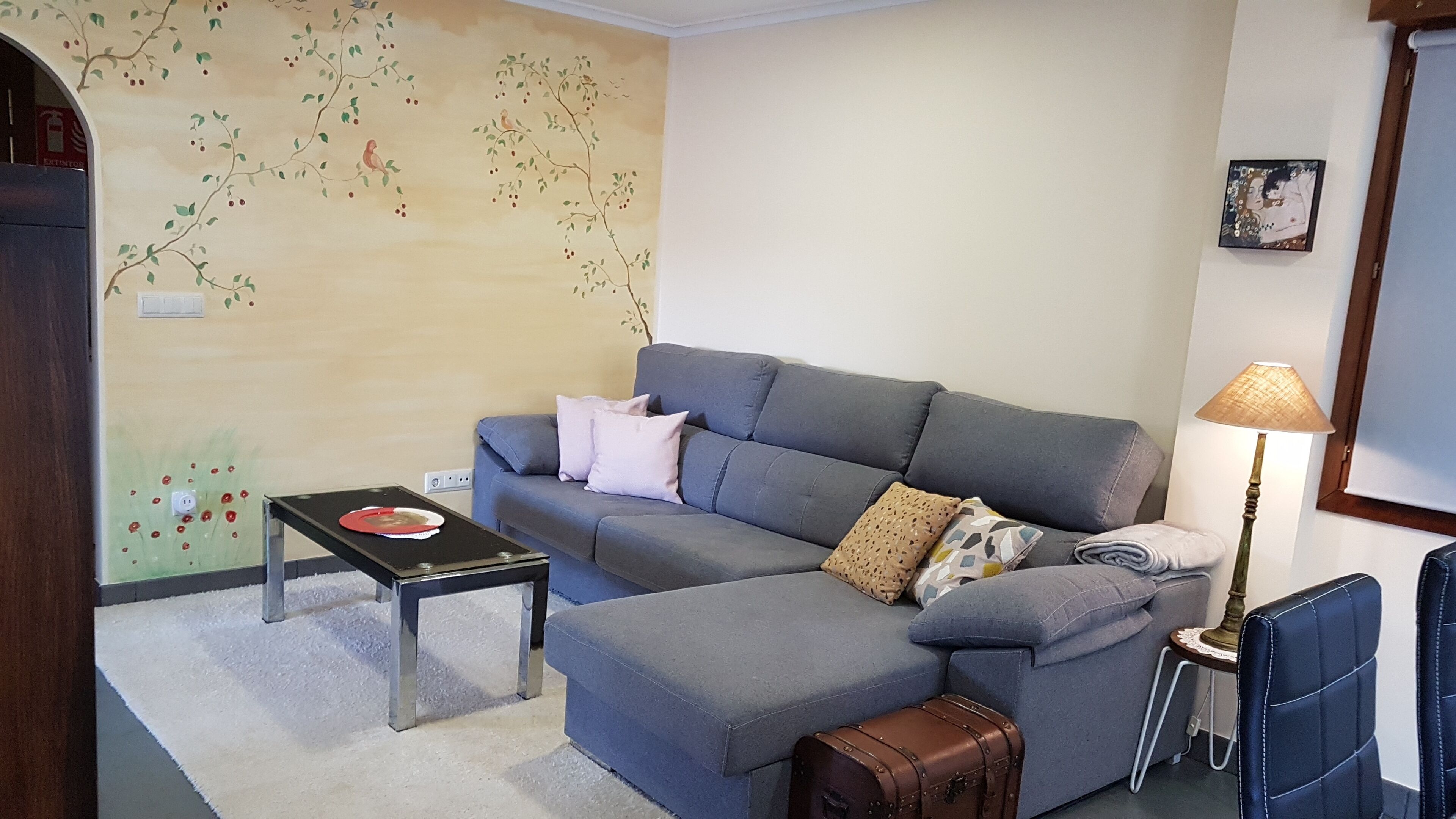 Een woonkamer met een grijze bank, een klein tafeltje, een lamp en een wand met een bloemenmuur.