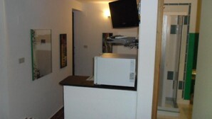 A small room with a white refrigerator, a microwave, and a flat-screen TV mounted on the wall. There is a door leading to a bathroom with a glass shower enclosure. - Townhome (Cefalù)