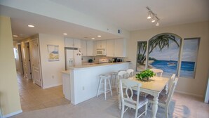 A modern kitchen with white cabinets, a central island, and bar stools. There is a dining table with chairs and a potted plant. The room has a large window with a view of the ocean and a palm tree.