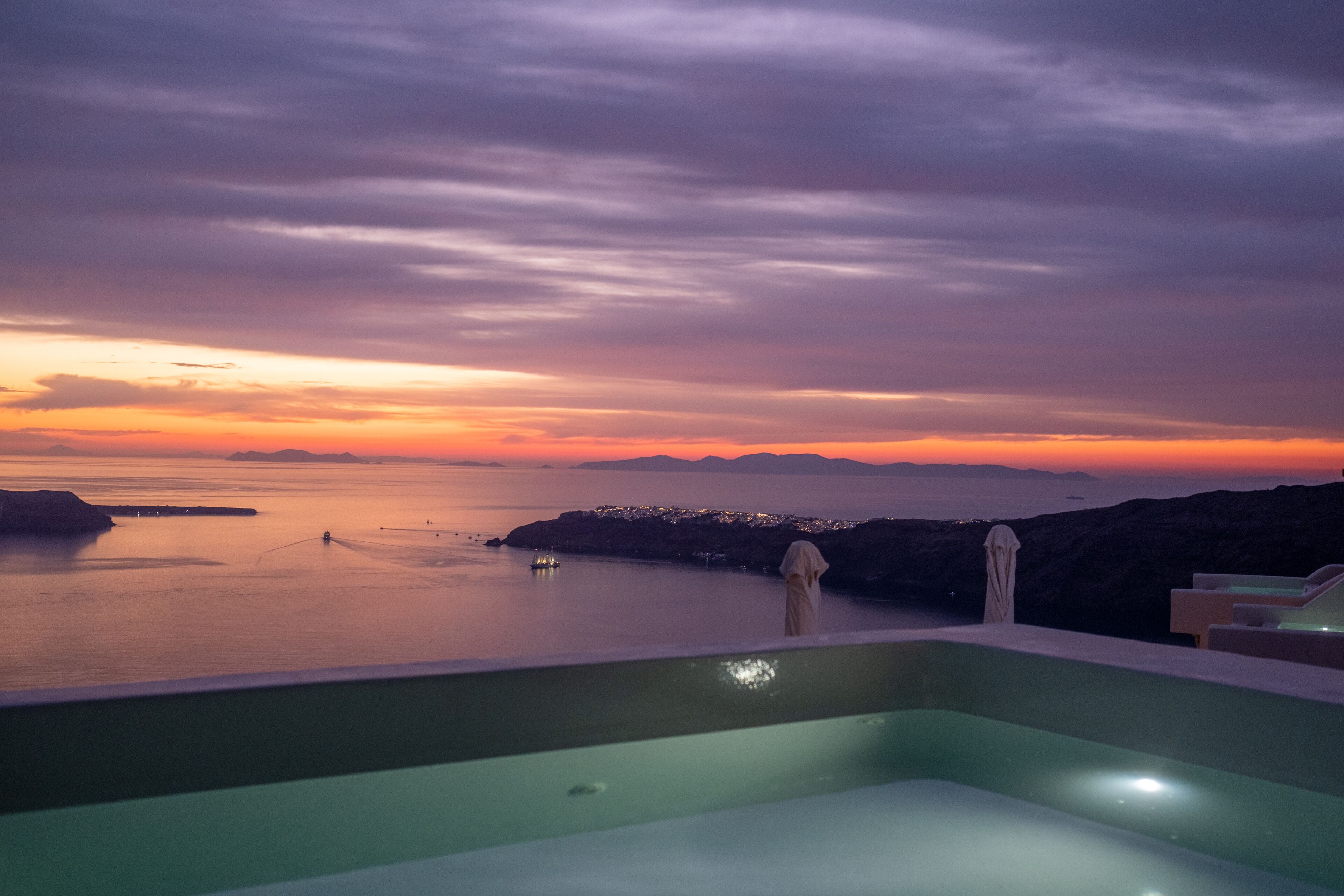 A sunset view from a balcony with a pool, overlooking a calm sea and distant land.