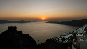 Vista costeira com um pôr do sol sobre o mar, edifícios com fachadas brancas e um primeiro plano rochoso.