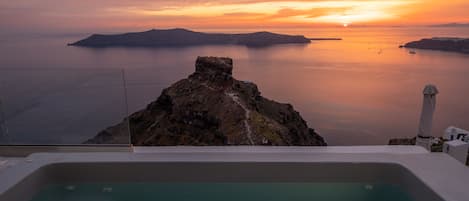 Une vue depuis un balcon, avec un jacuzzi, donnant sur un coucher de soleil au-dessus d’une mer calme et d’îles lointaines.