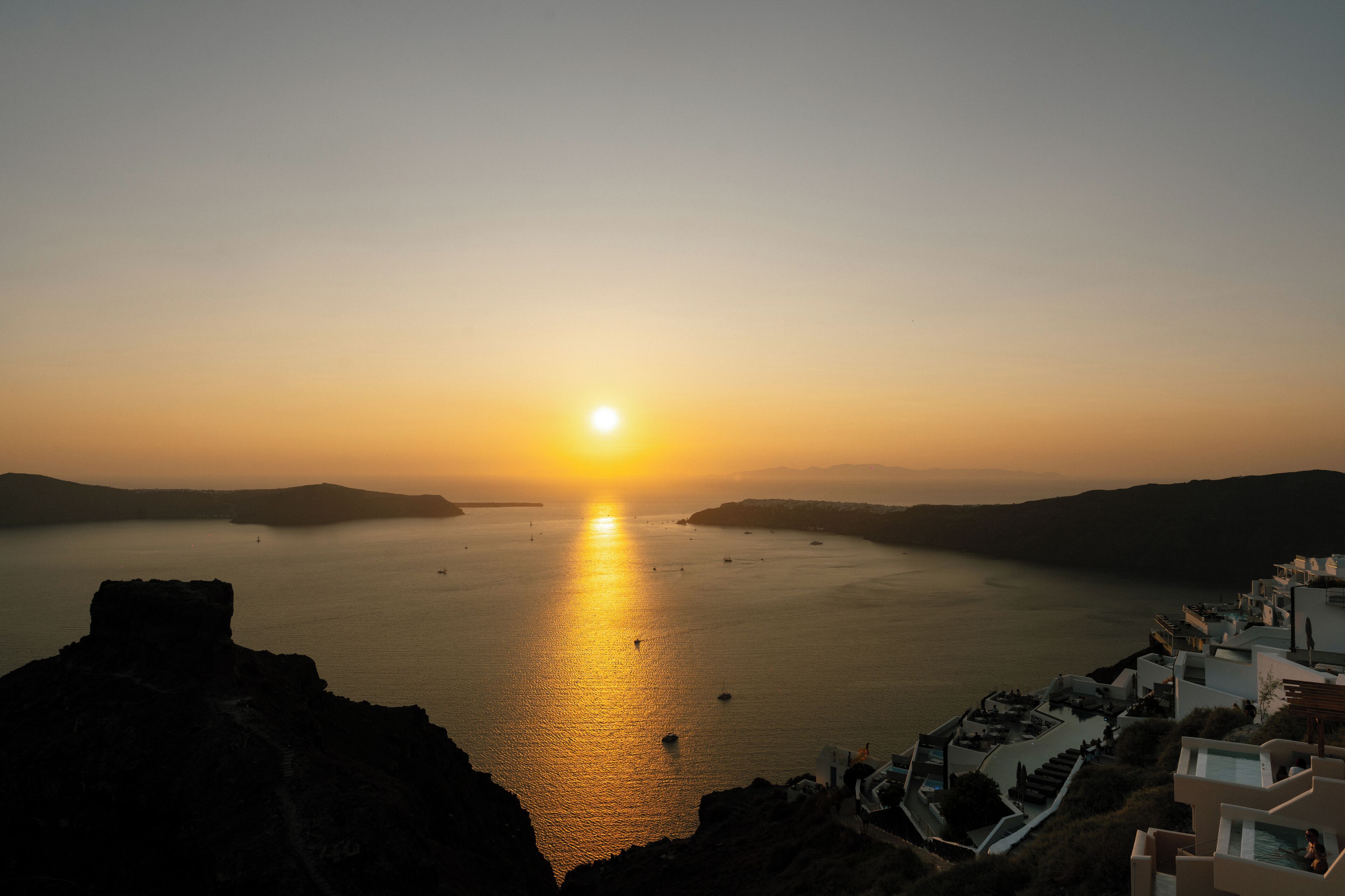 A coastal sunset view with a clear sky, a calm sea, and a white building with balconies overlooking the water.