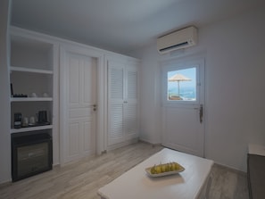A compact room with a white door, a window with an umbrella, a built-in air conditioner, and a small table with a fruit bowl. - Anemos Cave Suites (Santorini)