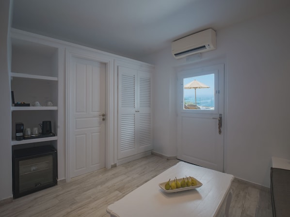 A compact room with a white door, a window with an umbrella, a built-in air conditioner, and a small table with a fruit bowl. - Anemos Cave Suites (Santorini)