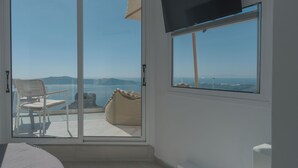 A room with a sliding glass door leading to a balcony with a chair and a bean bag. A flat-screen TV is mounted on the wall. - Anemos Cave Suites (Santorini)