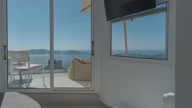 A room with a sliding glass door leading to a balcony with a chair and a bean bag. A flat-screen TV is mounted on the wall. - Anemos Cave Suites (Santorini)