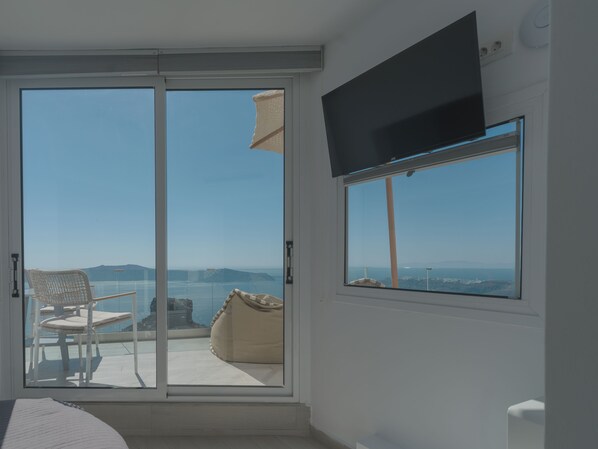 A room with a sliding glass door leading to a balcony with a chair and a bean bag. A flat-screen TV is mounted on the wall. - Anemos Cave Suites (Santorini)